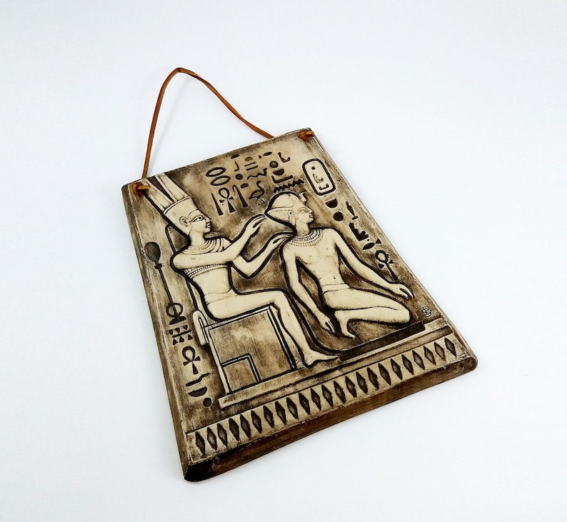K&ouml;nnte beinhalten: Eine braune Keramik-Wandplatte mit einem erhabenen Reliefdesign von zwei Figuren im alten &auml;gyptischen Stil. Die Figuren tragen traditionelle Kleidung und Kopfbedeckungen. Die Platte hat einen Seilhalter und ist mit Hieroglyphen verziert.