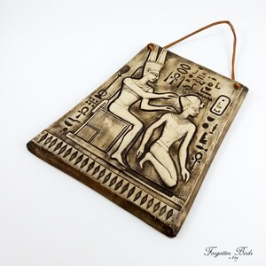 K&ouml;nnte beinhalten: Ein Wandbehang aus Keramik mit einer Reliefskulptur von zwei Figuren im Stil der alten &auml;gyptischen Kunst. Die Figuren sind in einer Szene dargestellt, in der ein Pharao einem knienden Menschen ein Geschenk darbringt. Der Wandbehang hat einen braunen Lederriemen zum Aufh&auml;ngen.