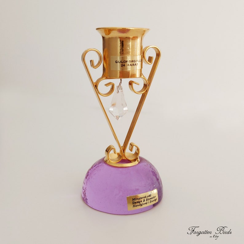 May include: A gold-plated metal candle holder with a purple glass base. The holder features a delicate, ornate design and a crystal drop hanging from the center. The base is inscribed with "M&aring;nsateras vid Design B. Bl&aring;mire Handgjord i Sverige".