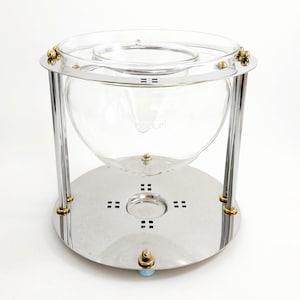 Peut inclure: Une cafetière en verre transparent et en chrome avec des détails dorés. La cafetière a une carafe en verre ronde et une base en métal avec de petits trous carrés. La marque "bodum" est visible sur le verre.