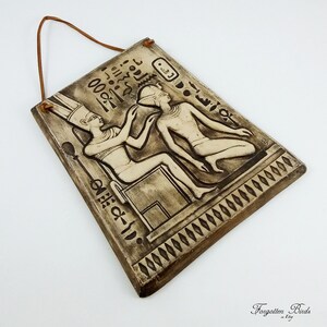 K&ouml;nnte beinhalten: Ein brauner Keramik-Wandbehang mit einer Reliefskulptur von zwei Figuren im alten &auml;gyptischen Stil. Die Figuren tragen aufwendige Kopfbedeckungen und Kleidung. Der Wandbehang hat eine Schlaufe aus Seil zum Aufh&auml;ngen und einen dekorativen Rand.