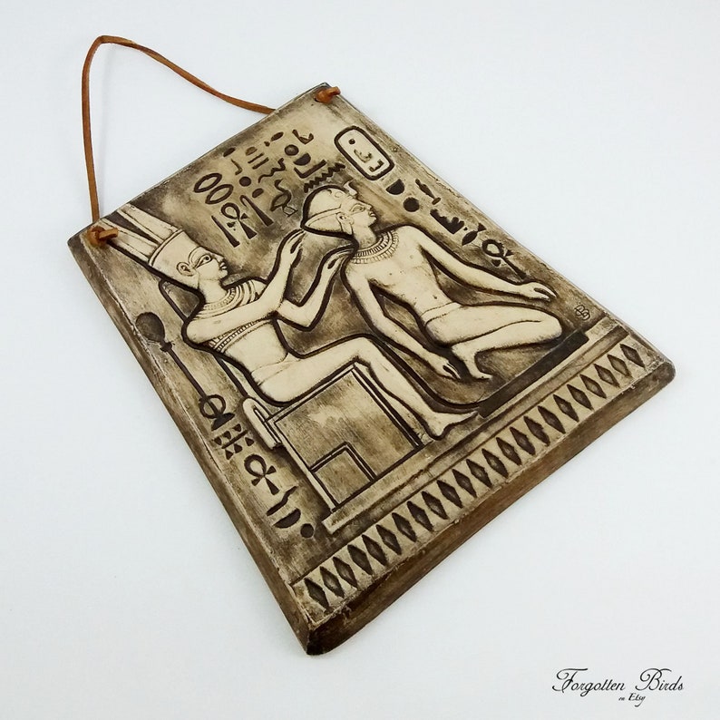 K&ouml;nnte beinhalten: Ein brauner Keramik-Wandbehang mit einer Reliefskulptur von zwei Figuren im alten &auml;gyptischen Stil. Die Figuren tragen aufwendige Kopfbedeckungen und Kleidung. Der Wandbehang hat eine Schlaufe aus Seil zum Aufh&auml;ngen und einen dekorativen Rand.