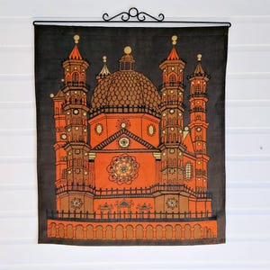 Puede incluir: Un gran tapiz de pared rectangular con una ilustración detallada de una catedral o mezquita en tonos naranja, marrón y beige sobre un fondo gris oscuro. La obra de arte está montada en una barra de metal negro con adornos decorativos.