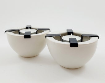 以下が含まれることがあります： 黒い金属製のキャンドルホルダーが付いた白いセラミック製のキャンドルホルダー2つ。各ホルダーは丸い形をしていて、黒い金属製のフレームに4本の腕が付いています。