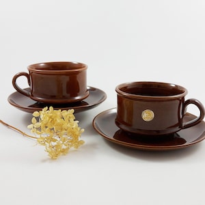 Könnte beinhalten: Zwei braune Keramik-Teetassen mit passenden Untertassen. Die Tassen haben einen goldenen Aufkleber an der Seite. Die Tassen stehen auf einer weißen Oberfläche mit einem kleinen Strauß getrockneter Blumen.