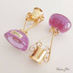 May include: Two purple glass bottle stoppers with gold metal stands. The stoppers are round and have a smooth, shiny surface. The stands are ornate and have a delicate, intricate design. The stoppers are labeled "Murrhudda Design by Blomster Handgjord Sverige".
