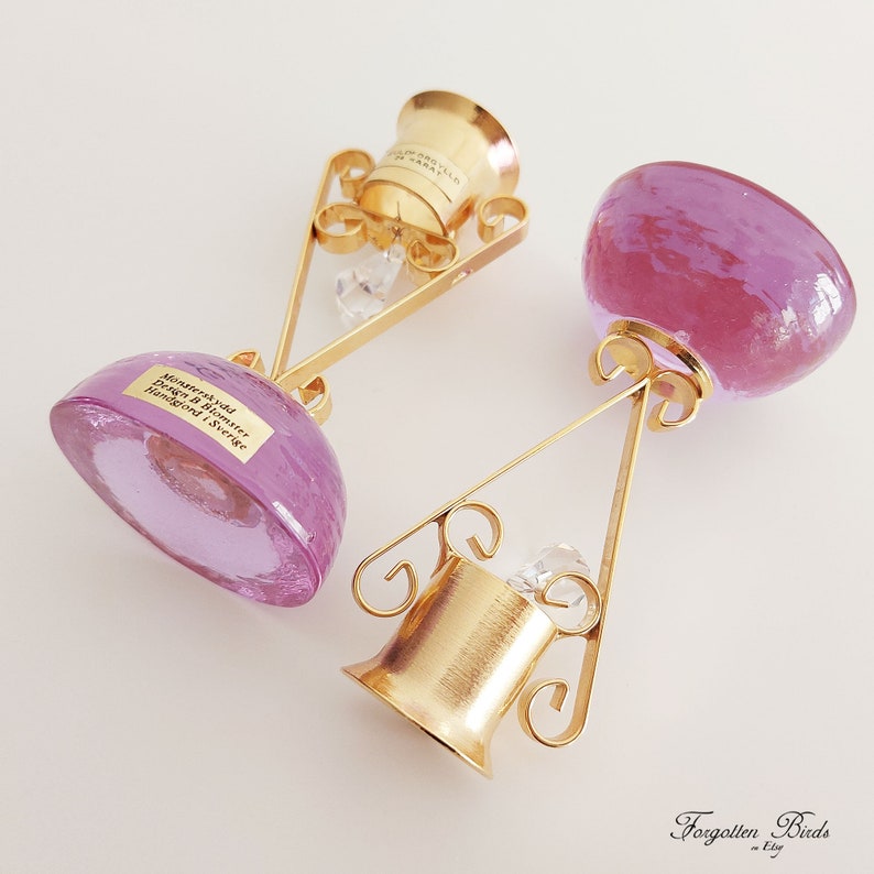 May include: Two purple glass bottle stoppers with gold metal stands. The stoppers are round and have a smooth, shiny surface. The stands are ornate and have a delicate, intricate design. The stoppers are labeled "Murrhudda Design by Blomster Handgjord Sverige".