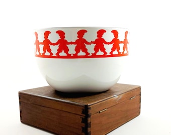 May include: A white ceramic bowl with a red band of stylized figures holding hands. The bowl sits on a small, dark wooden box with visible dovetail joints. The background is plain white.