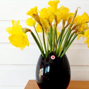 May include: A black, glossy vase holding a bouquet of bright yellow daffodils. The vase has a unique, egg-like shape with a flared top. The flowers have green stems and are in full bloom, creating a vibrant contrast against the dark vase.