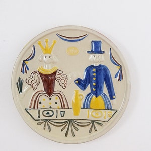 May include: A ceramic plate with a stylized illustration of a king and queen in traditional clothing. The plate is decorated with yellow, blue, and brown accents. The text "Villa" is visible in the center of the plate.