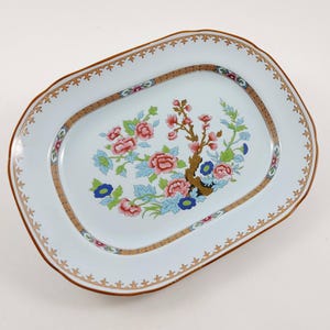 Op de afbeelding: Ovale serveerschaal met een witte achtergrond en een bloemenontwerp. De schaal heeft een bruine rand en een decoratieve goud-bruine binnenrand. Het bloemenontwerp toont roze en blauwe bloemen met groene bladeren.