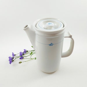 May include: White ceramic coffee pot with a blue floral design and a lid. The pot has a handle and a spout. A bunch of purple flowers is in the background.