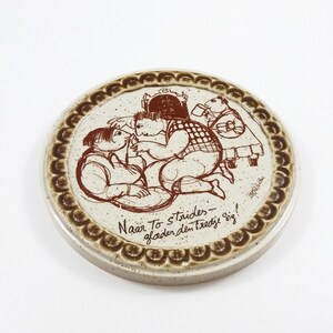 May include: A brown and white ceramic coaster with a brown illustration of three people sitting at a table. The text "Naar To strijden - gladden den tredje sig!" is written in brown cursive on the coaster.