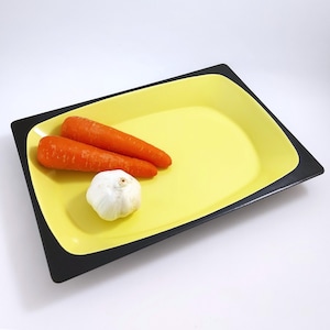 Könnte beinhalten: Eine rechteckige gelbe Servierplatte mit schwarzem Rand, auf der zwei orange Karotten und eine weiße Knoblauchzehe liegen.