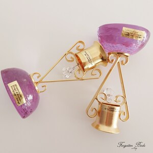 May include: Two gold-colored bottle stoppers with crystal accents, each with a purple glass base. The stoppers are labeled "GULOF ORGYLLD 24 KARAT" and "FORGYLLD KARAT".