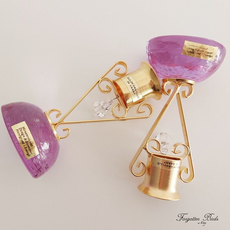 May include: Two gold-colored bottle stoppers with crystal accents, each with a purple glass base. The stoppers are labeled "GULOF ORGYLLD 24 KARAT" and "FORGYLLD KARAT".