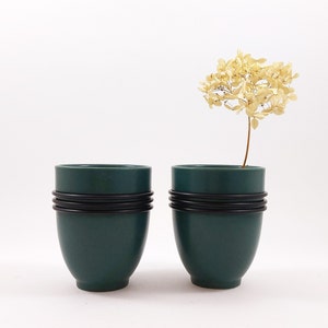 Op de afbeelding: Twee donkergroene keramische vazen met zwarte banden rond het midden. De vazen staan op een wit oppervlak. Een gedroogde witte bloem staat in de vaas rechts.