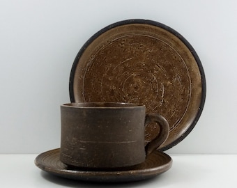 May include: A brown ceramic teacup and saucer set with a textured, swirling design. The cup has a handle and the saucer has a slightly raised edge.