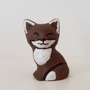 May include: A brown ceramic figurine of a cat with white markings on its face, chest, and paws. The cat is sitting with its eyes closed and a smile on its face.