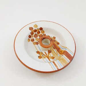 May include: A white ceramic candle holder with a brown and blue floral design. The holder has a round base and a raised center for a candle.
