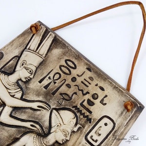 K&ouml;nnte beinhalten: Ein Keramik-Wandbild mit einer Reliefskulptur von zwei &auml;gyptischen Figuren. Die Figuren tragen traditionelle Kopfbedeckungen und Schmuck. Der Hintergrund ist mit Hieroglyphen und anderen &auml;gyptischen Symbolen verziert.