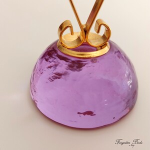 May include: A purple glass base with a gold metal ring and a decorative swirl design. The base is round and has a smooth, polished surface.