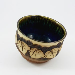 Pode incluir: Uma tigela pequena de cerâmica marrom com um esmalte azul e verde no interior. O exterior da tigela é decorado com um padrão de folhas marrons.