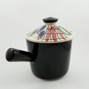 May include: A black ceramic pot with a white lid featuring a colorful geometric design. The pot has a single handle and is perfect for serving a small meal.