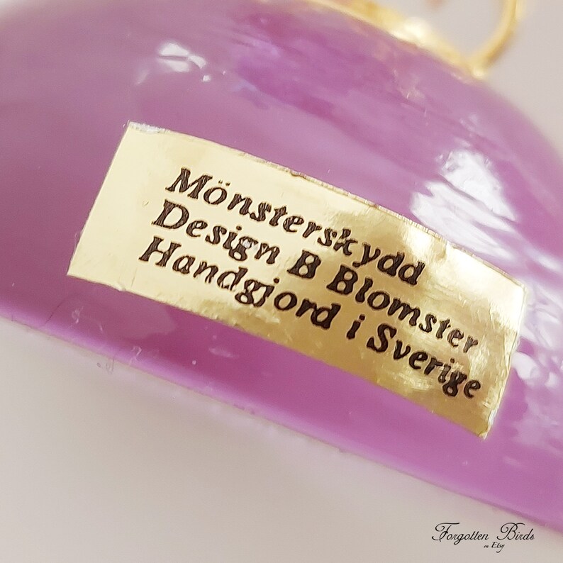 May include: A purple glass ornament with a gold label that reads "M&ouml;nsterskydd Design B Blomster Handgjord i Sverige".