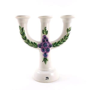 May include: A white ceramic candelabra with three arms. The candelabra is decorated with green leaves and purple and blue floral accents. The base is round and the arms are curved.