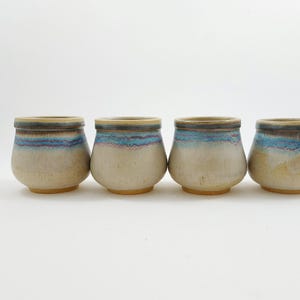 Könnte beinhalten: Set aus vier Keramikbechern mit hellbeigem Überzug und blauen und braunen Akzenten. Die Becher haben eine abgerundete Form und eine leicht ausgestellte Lippe.