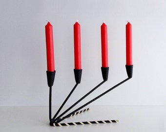 May include: A black metal candle holder with four arms, each holding a red candle. The holder has a decorative base made of black and white striped paper.