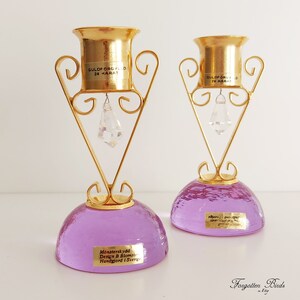 May include: Two gold-plated candle holders with purple glass bases. Each holder features a crystal drop and a label that reads "Guldforgyld 24 Karat". The base of each holder has a label that reads "M&ouml;nsterskydd Design B Blomster Handgjord i Sverige".