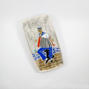 May include: A ceramic dish with a white background and a hand-painted design of a sailor playing an accordion. The sailor is wearing a blue and white striped shirt and a blue hat. The dish is decorated with gold accents and a blue and white wave pattern.
