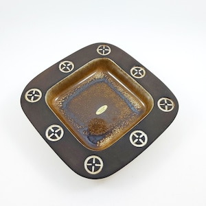 May include: A square, brown ceramic dish with a lighter brown interior. The rim is dark brown with white cross-like designs. The interior has a speckled, glossy finish. A small sticker is visible inside the dish.