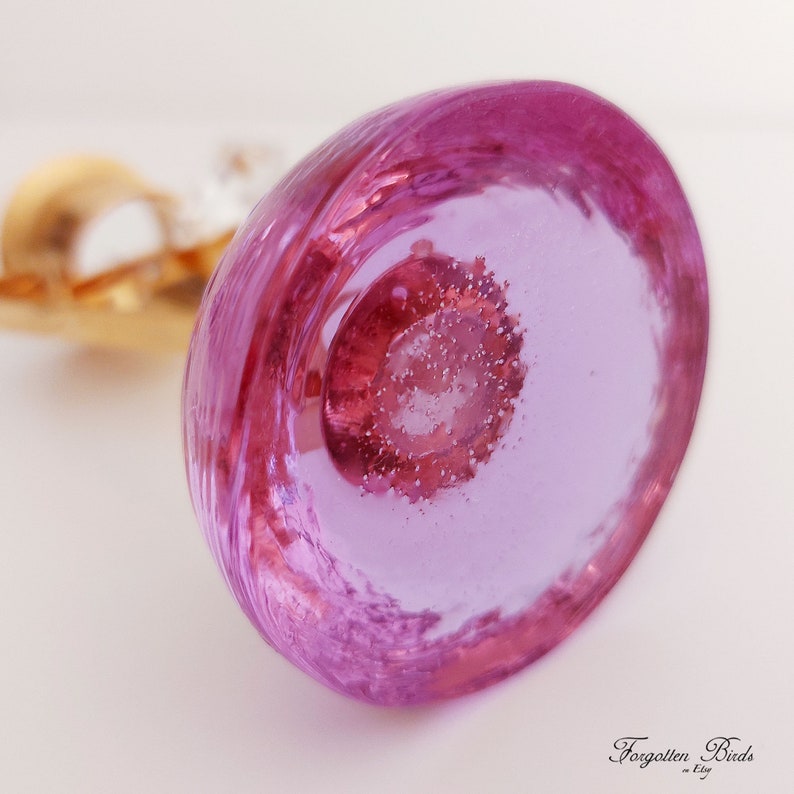 May include: A close-up of a pink glass bottle stopper with a clear center and a swirling pattern. The stopper has a small, clear, round, textured piece in the center.