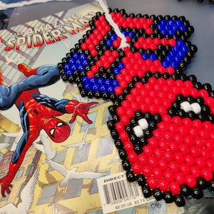 May include: A red, blue, and black beaded Spiderman figure. The figure is made of small, round beads and is shaped like Spiderman's head and torso. The figure is hanging from a white string.