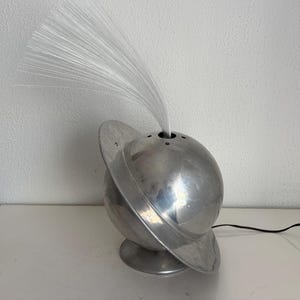 May include: A silver-colored fiber optic lamp with a spherical base and a ring around the middle. The lamp emits a cluster of white fiber optic strands from the top. The lamp is on a white surface.