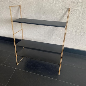 May include: A gold metal shelf unit with three black shelves. The shelves are made of wood and have a smooth finish.