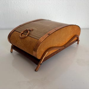 Puede incluir: Una caja de almacenamiento rectangular con una tapa de madera curvada de listones y un asa de anillo de metal. Los lados están cubiertos con un material beige texturizado y toda la estructura se apoya en un marco de metal cobrizo con cuatro patas.