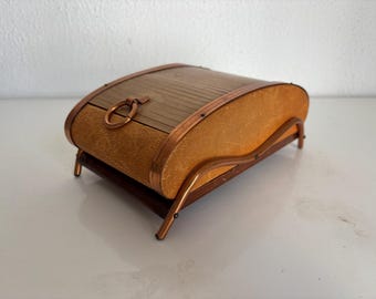 Peut inclure: Une boîte de rangement rectangulaire dotée d'un couvercle en bois incurvé à lattes avec une poignée en anneau métallique. Les côtés sont recouverts d'un matériau beige texturé et l'ensemble repose sur un cadre en métal cuivré à quatre pieds.