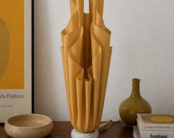 May include: A tall, mustard yellow table lamp with a sculptural, pleated shade that mimics the appearance of folded fabric. The lamp is supported by a circular white marble base and includes a power cord extending from the bottom side.