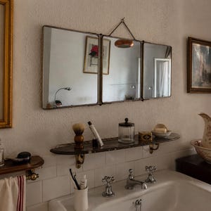 May include: A triptych wall mirror featuring three rectangular glass panels with thin dark metal frames, suspended by a metal chain. The mirror reflects a room interior and is positioned above a dark marble shelf holding a shaving brush.