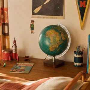 Puede incluir: Un escritorio de estilo vintage con un globo terráqueo verde sobre una base de madera oscura, un cohete de juguete de metal rojo, un cómic Le Journal de Spirou, un portalápices azul con triángulos blancos y libros coloridos sobre madera.