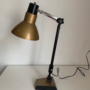 May include: A gold and black adjustable desk lamp with a black cord. The lamp has a round, gold-coloured shade and a black base with a knob.