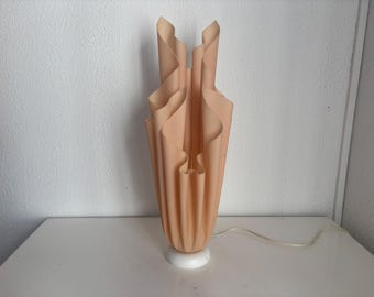 May include: A tall table lamp featuring a peach-colored, pleated shade with a vertical, undulating design that flares outward at the top. The shade is supported by a circular white base and includes a thin, white electrical cord extending outward.