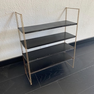 May include: A vintage-style, four-tiered shelf unit. The shelves are black, and the frame is a gold color. The shelves are supported by a gold-colored metal frame. The unit is designed to stand on the floor and is suitable for displaying items.