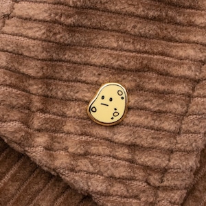 Może przedstawiać: Złota przypinka z kreskówkowym wzorem ziemniaka. Przypinka ma jasnożółte tło z czarnymi rysami twarzy i małymi czarnymi kropkami. Przypinka jest umieszczona na brązowej tkaninie sztruksowej.