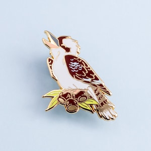 Op de afbeelding: Goudkleurige metalen broche met een kookaburra vogel op een tak met bessen. De vogel heeft witte, bruine en crèmekleurige veren. De broche heeft een glanzende afwerking en staat tegen een lichtblauwe achtergrond.