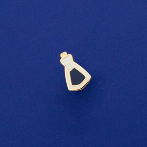May include: A gold-coloured enamel pin shaped like a bottle with a white outline and a dark blue fill.